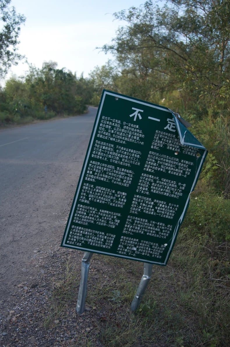 MOSS-Image: A weathered green roadside sign covered in small Chinese text, standing at the edge of a country road at dusk